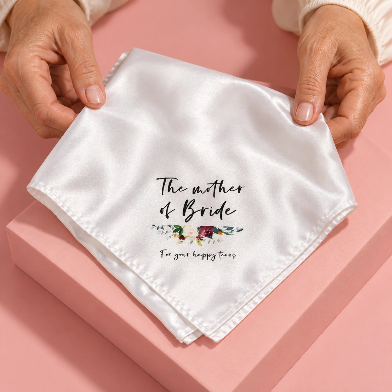 A white satin square handkerchief with black script text and a small floral illustration, held by hands over a pink gift box. The text reads 'The mother of Bride For your happy times'.