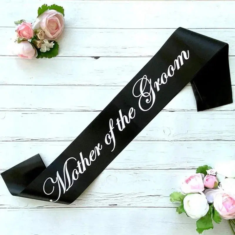 Black sash with 'Mother of the Groom' text on a white wooden background with floral decorations.