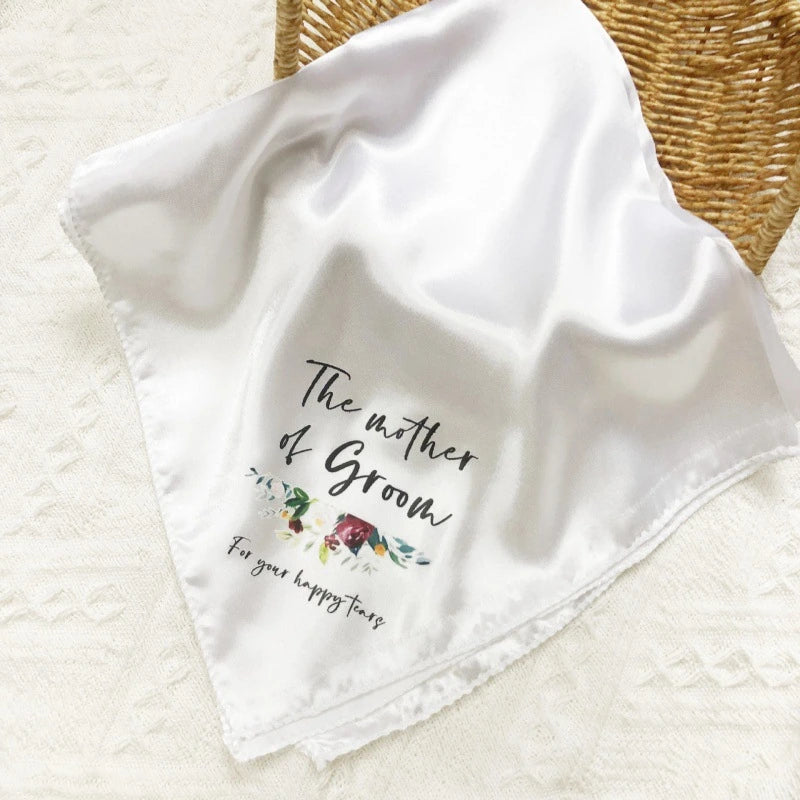 A white satin baby bandana with 'The mother of Groom' and 'For your happiness first' printed in black script with a small floral illustration, draped on a textured white fabric next to a woven basket.