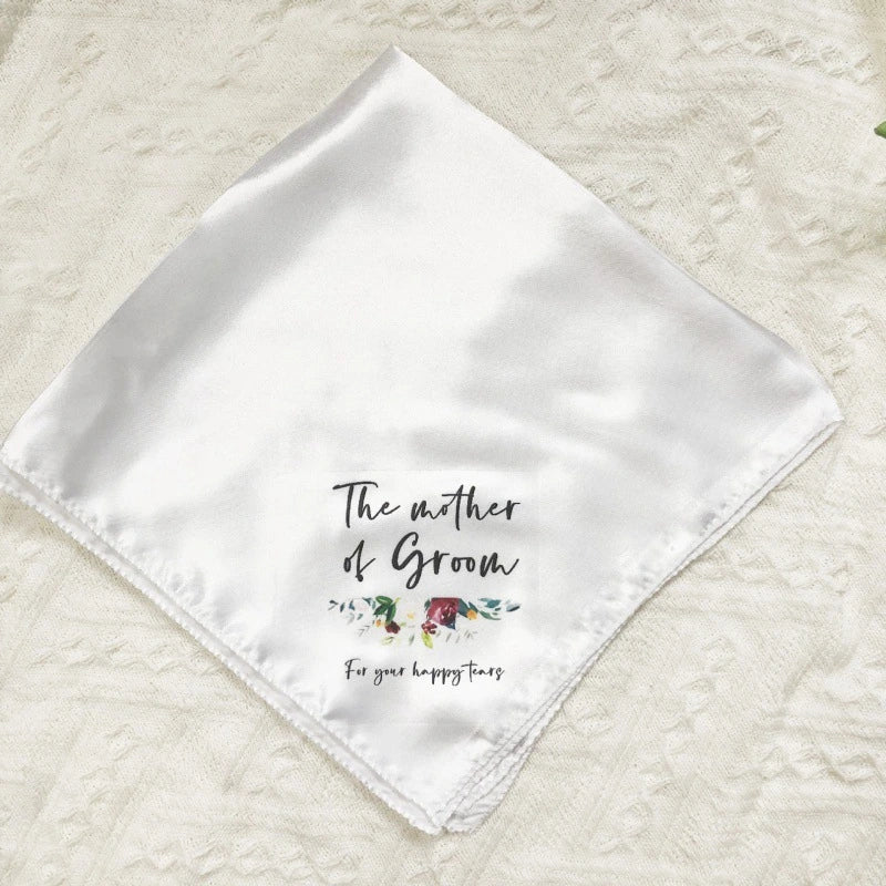 A white satin square bandana gift wrap featuring the printed text 'The mother of Groom' and 'For your happy groom' with a small floral illustration, laid flat on a textured cream fabric background.