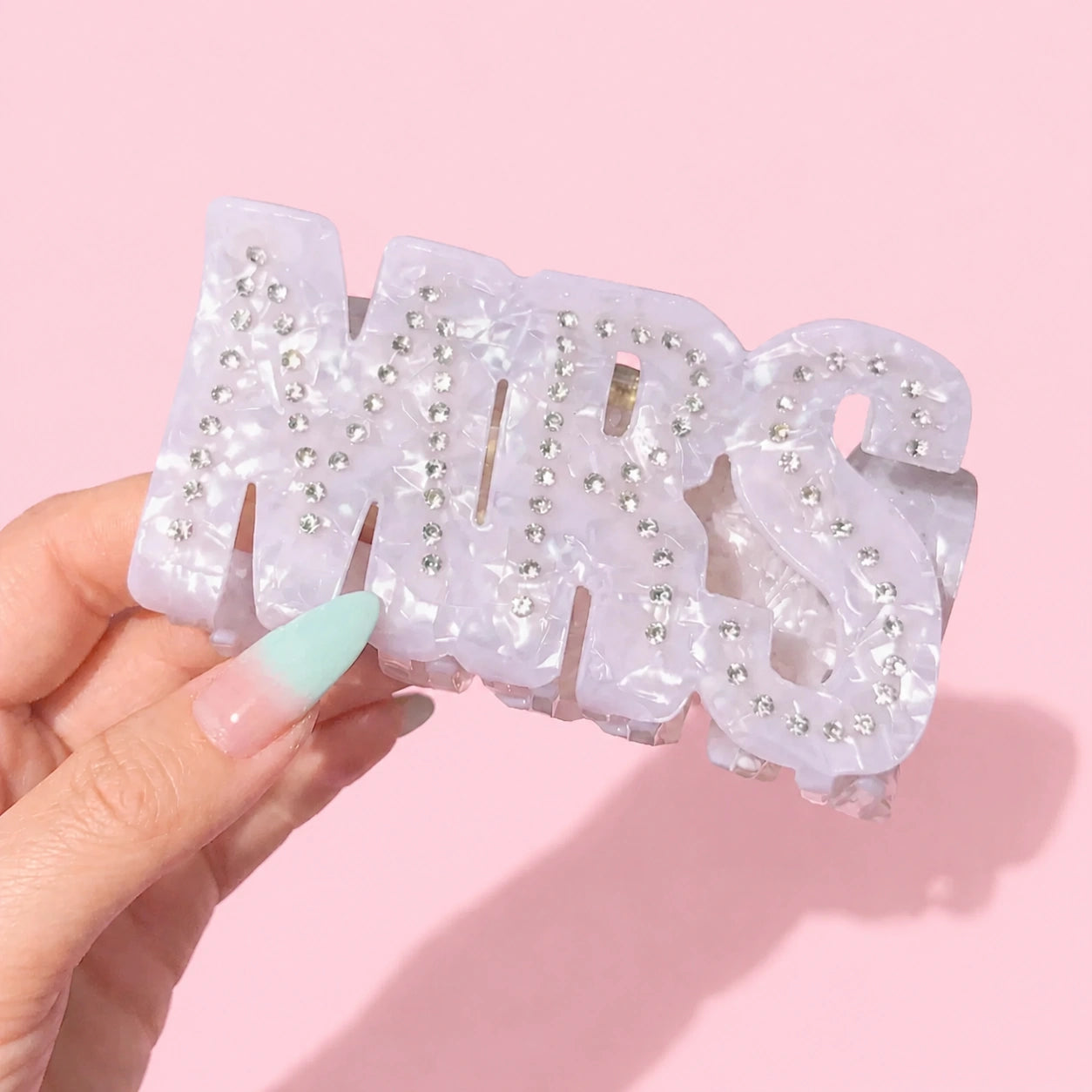 Hand holding a 'MRS' hair clip with rhinestones against a pink background