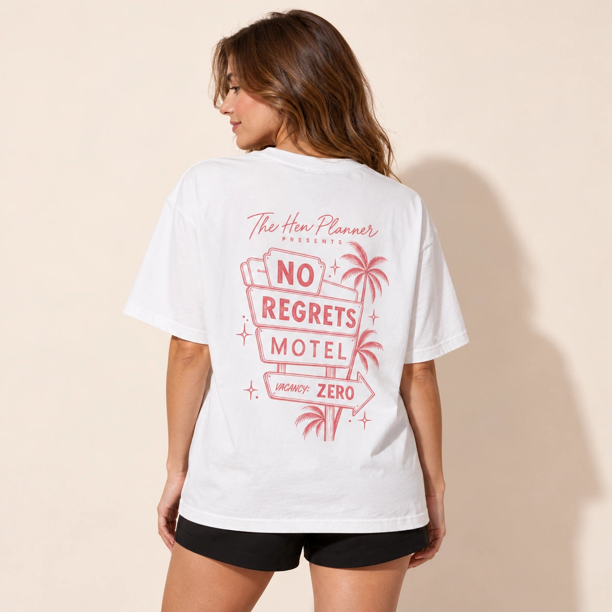 A woman shown from the back wearing a white graphic T-shirt with a pink retro-style 'No Regrets Motel' design and black shorts, standing against a plain beige wall.