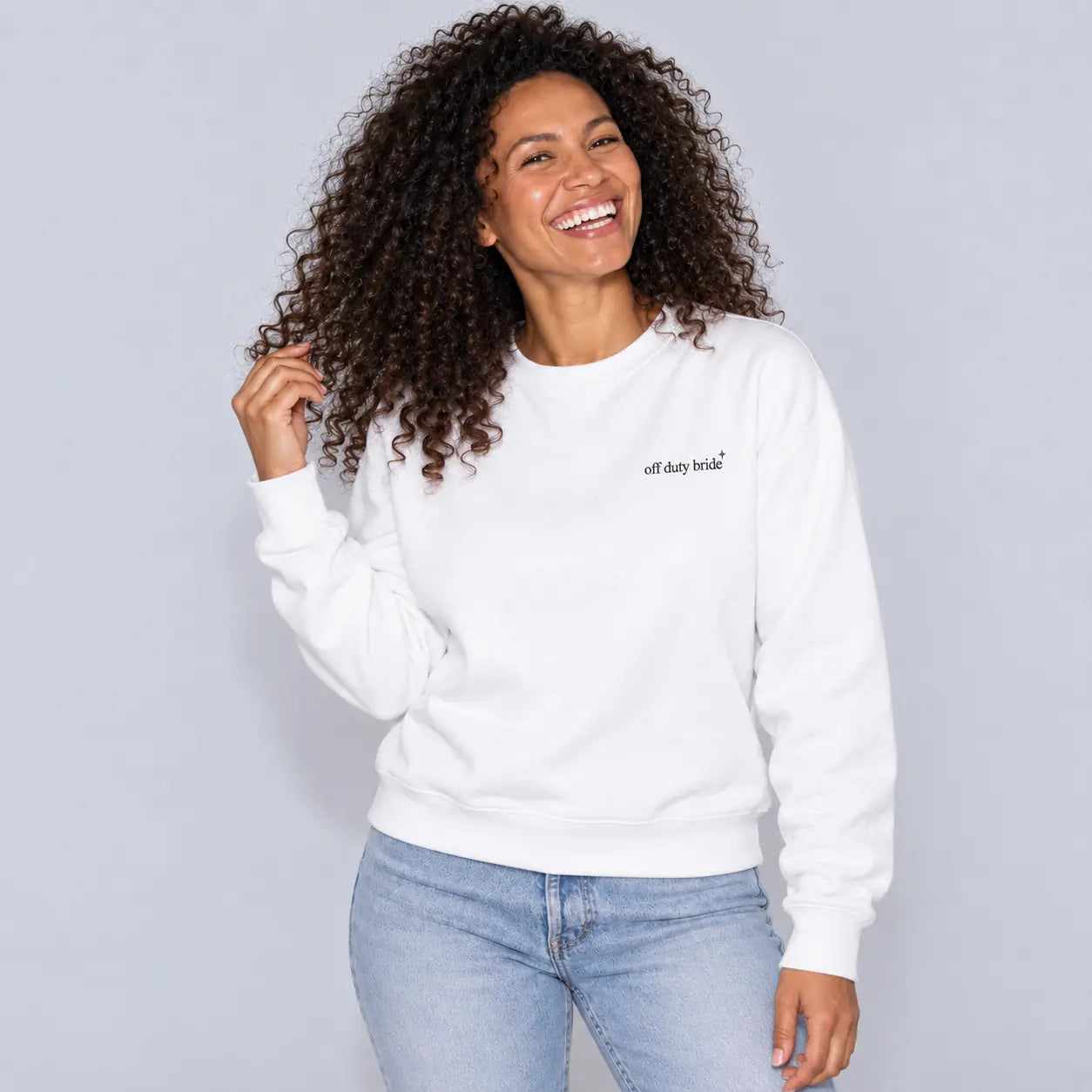 A woman with curly dark hair smiling while wearing a white crewneck sweatshirt with 'off duty bride' embroidered in black.