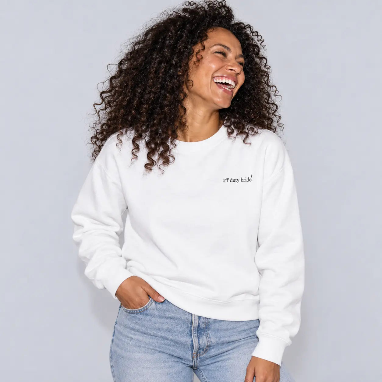 A woman with curly dark hair is smiling and laughing while wearing a white crewneck sweatshirt with 'off duty bride' .