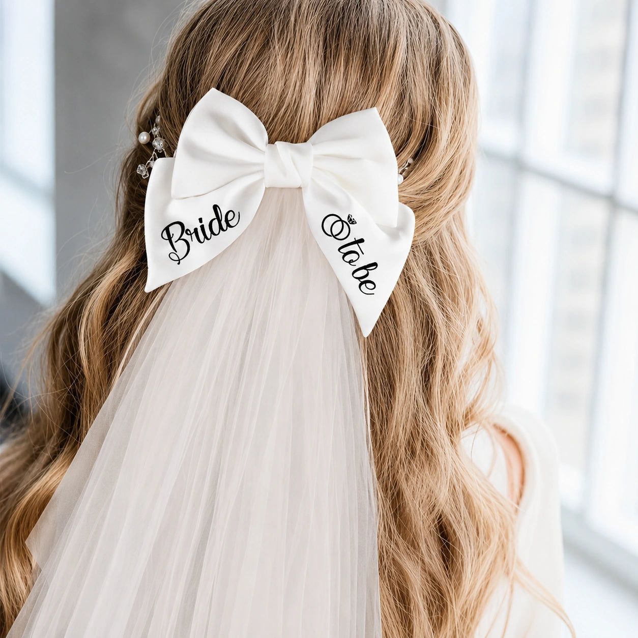 A white bridal hair bow hair clip with 'Bride to be' embroidered in black script, worn on a woman's blonde hair with a tulle veil.