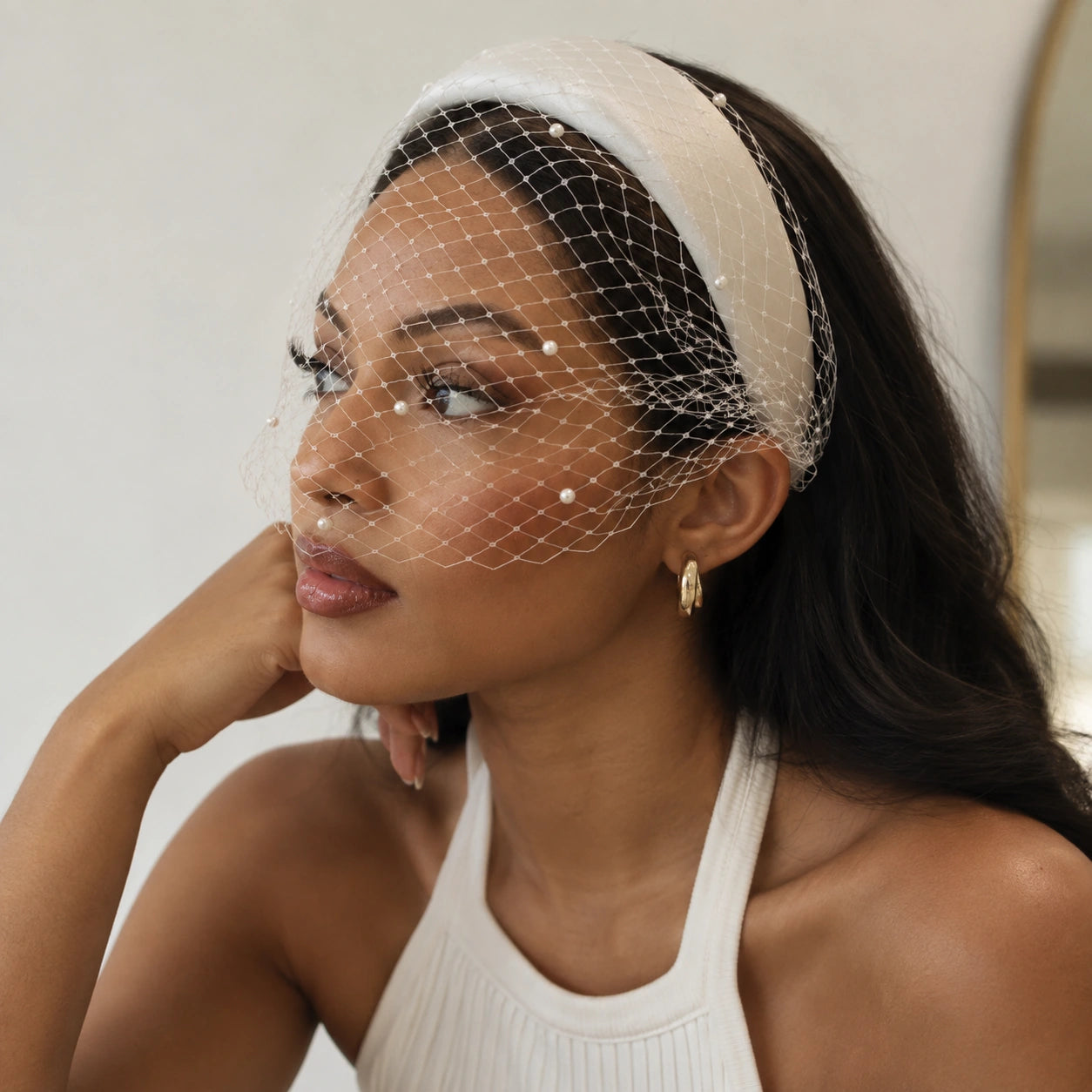 A woman wearing a white fabric headband with a delicate white net veil and small pearl-like embellishments, styled with a white ribbed halter top.