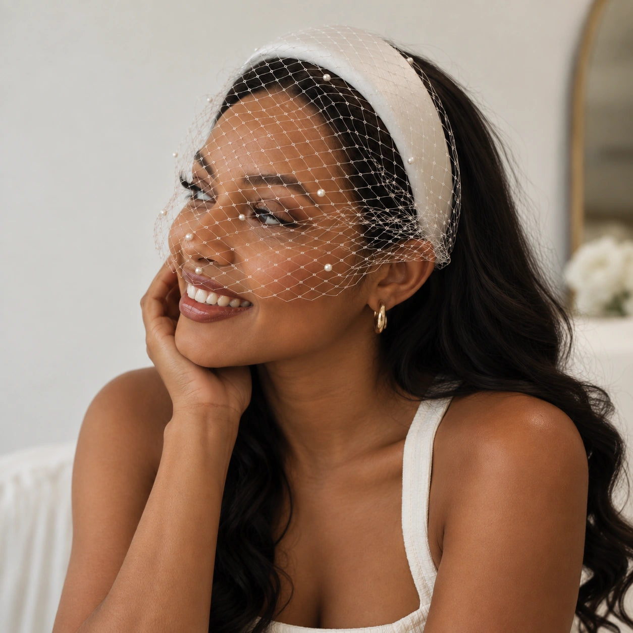 A woman wearing a white bridal veil with delicate pearl embellishments and a matching white headband, posing in a softly lit indoor setting.