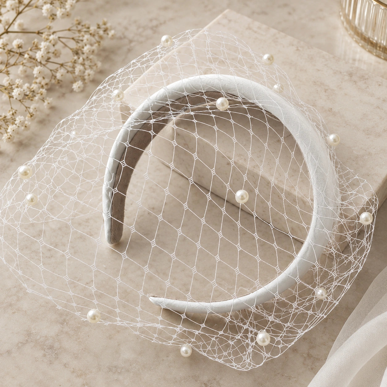 A white bridal veil with pearl accents draped over a white headband, styled on a beige marble surface.