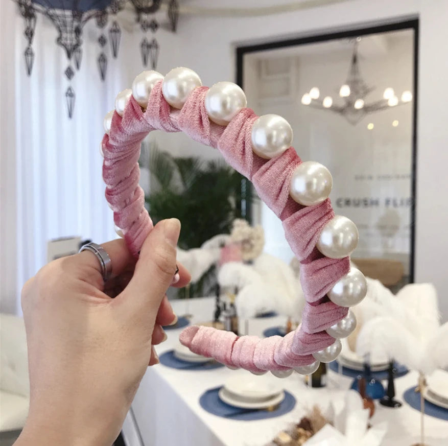 A hand holds a pink velvet headband decorated with white faux pearls, set against a blurred indoor background of a decorated table and mirror.