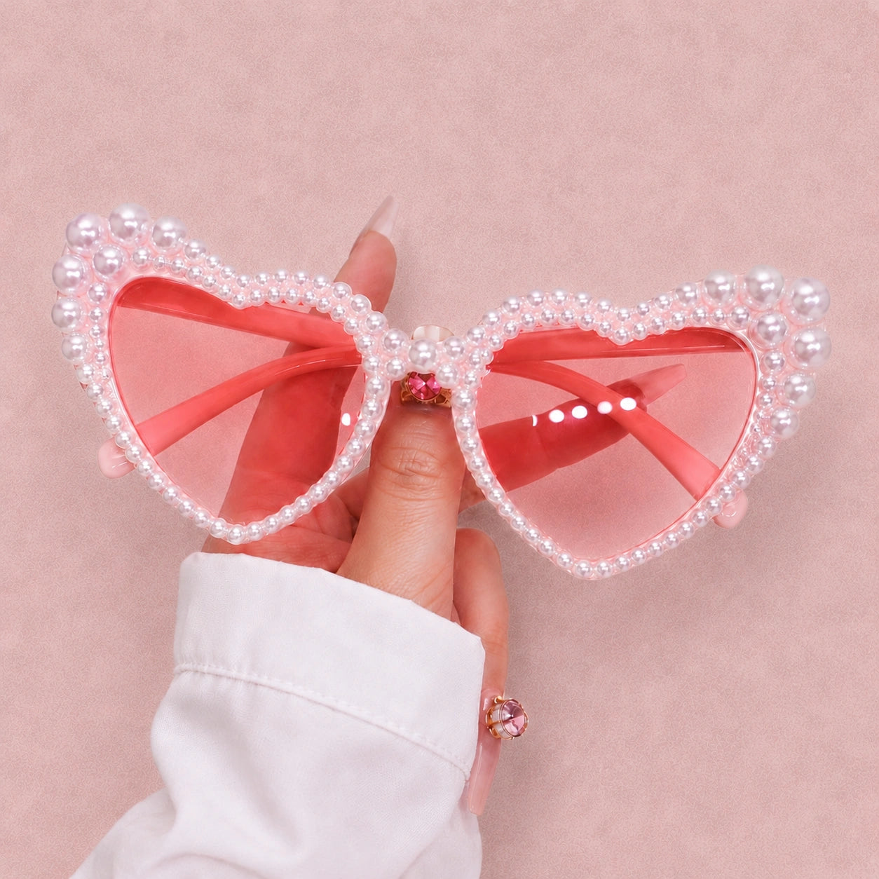 Heart-shaped sunglasses with pearl details held by a hand against a pink background