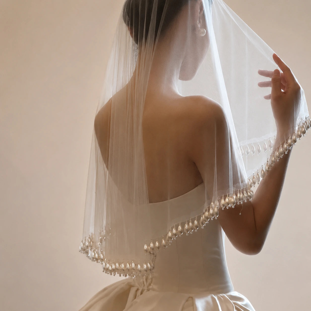 Woman wearing a white wedding dress with a pearl-trimmed veil against a neutral background