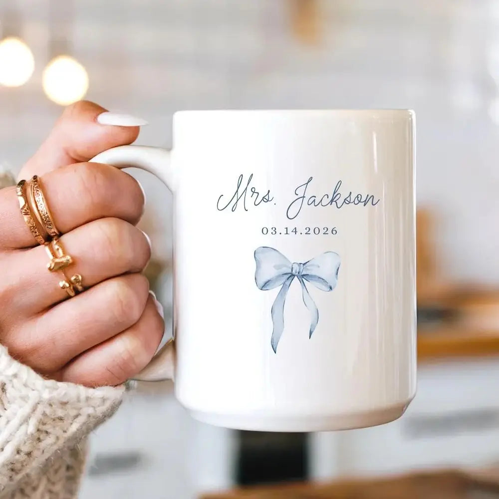 White mug with a blue bow design and personalized text held by a hand.