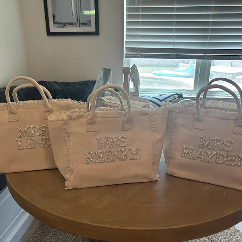 Three beige tote bags with embroidered names on a wooden table.