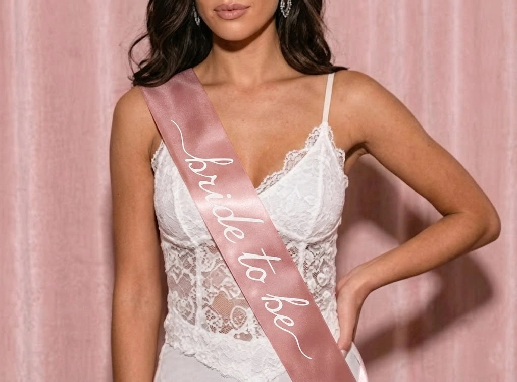 A woman wearing a white lace bodysuit and a dusty pink satin sash that reads 'bride to be' poses against a pink curtain backdrop.