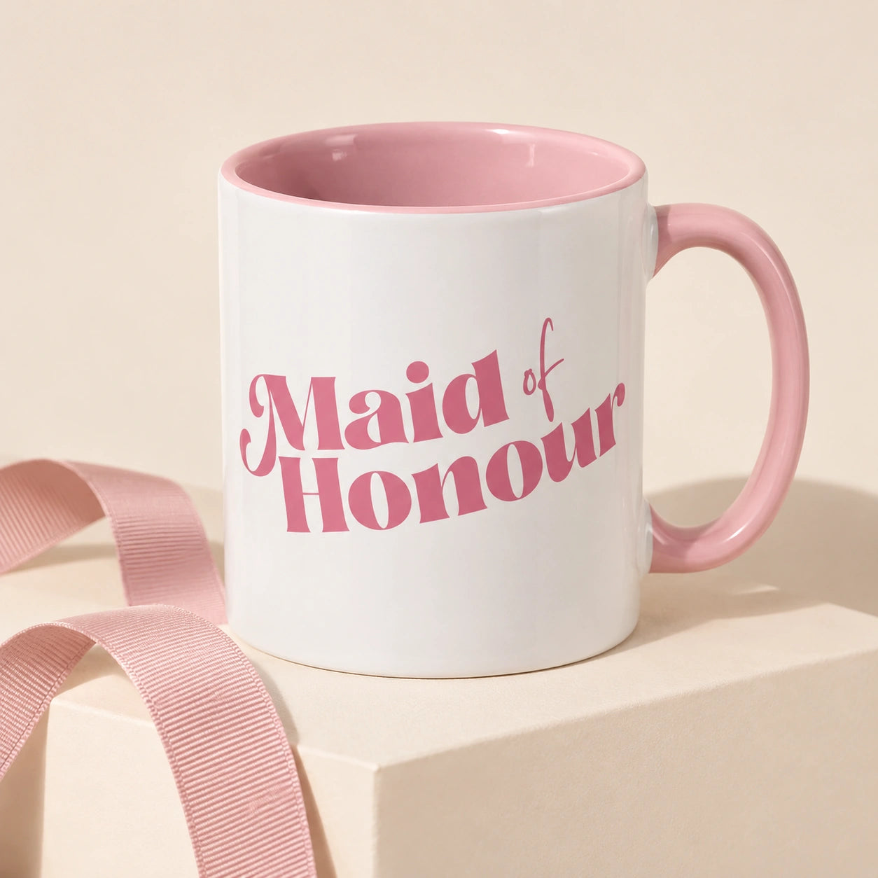 A white ceramic mug with a pink interior and handle, printed with the words 'Maid of Honour' in pink script, displayed on a cream pedestal with a pink ribbon.