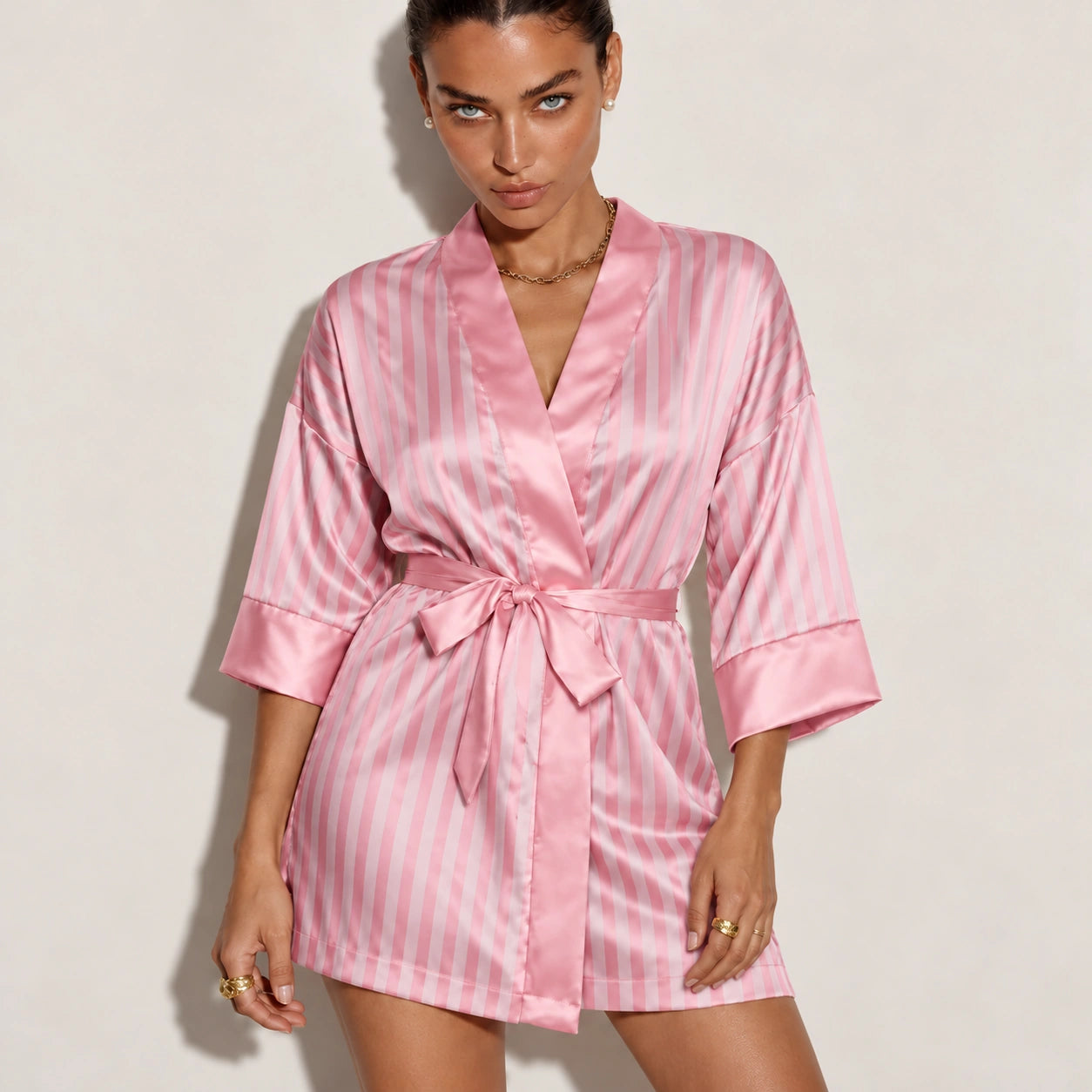 A woman modeling a pink satin robe with vertical light pink and white stripes, featuring a tie waist and elbow-length sleeves, against a plain light beige background.