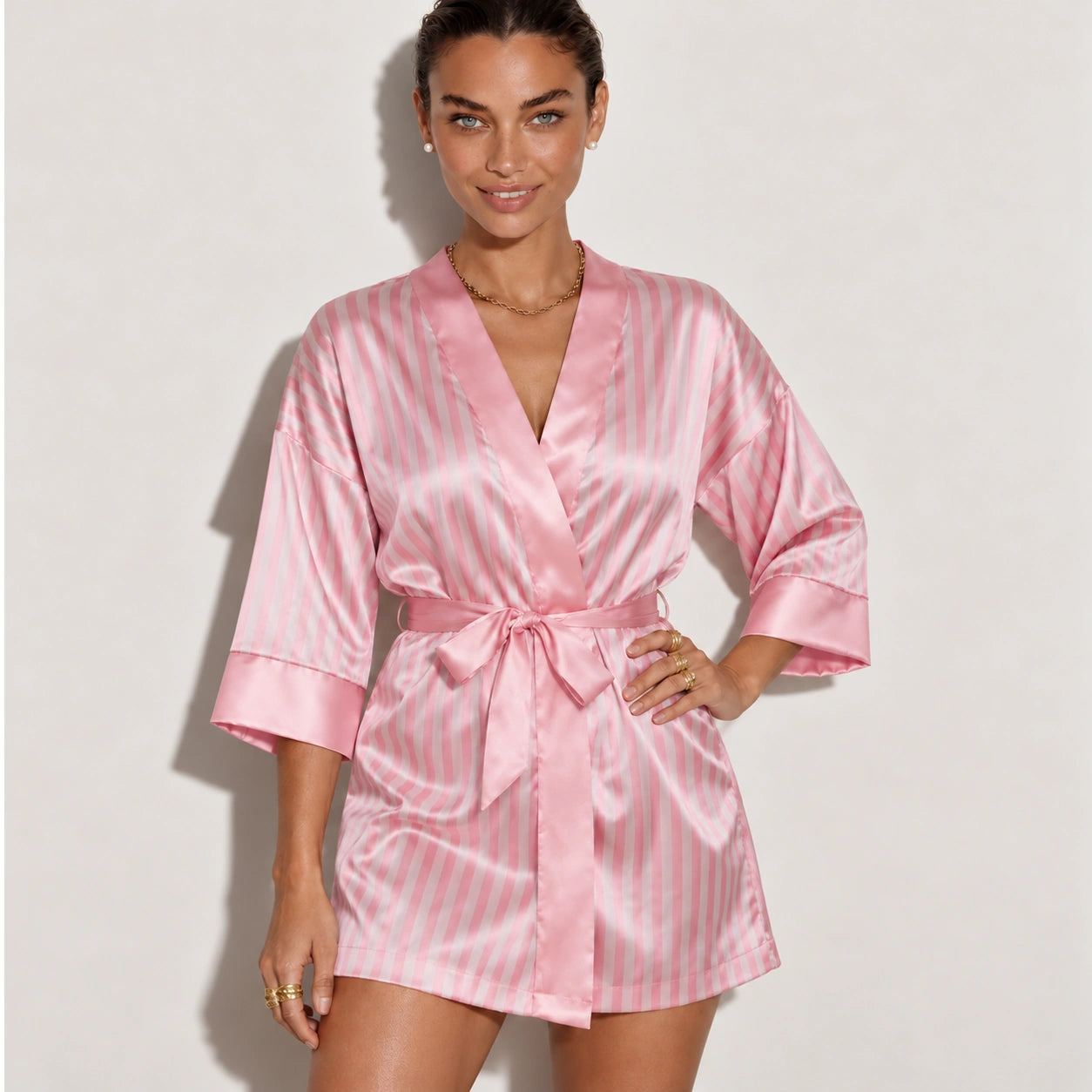 A woman modeling a pink satin robe with thin white vertical stripes, featuring a tie waist belt and three-quarter sleeves, set against a plain light gray studio background.