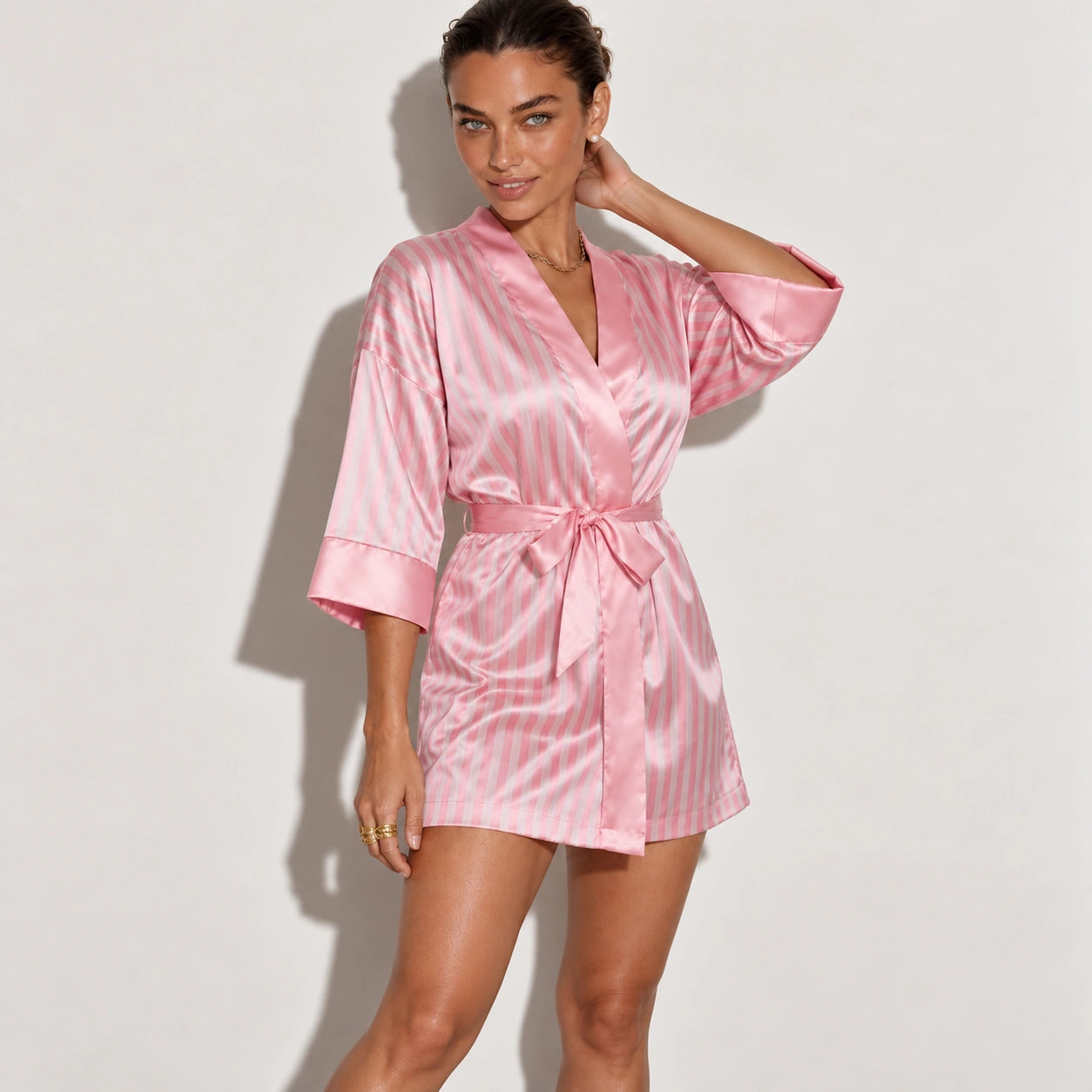 A woman modeling a pink satin robe with vertical white stripes, featuring a tie waist belt and elbow-length sleeves.