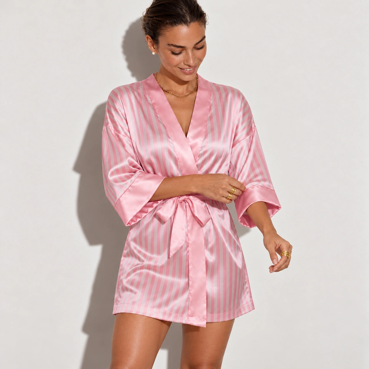 A woman wearing a pink satin robe with thin white vertical stripes, standing against a plain light gray wall.