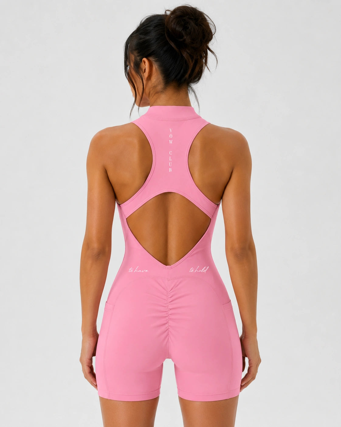 A woman shown from the back wearing a pink athletic one-piece jumpsuit with a high neck and a large open cutout at the back, photographed against a light gray studio background.