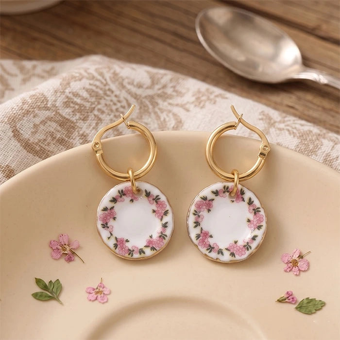 A pair of gold hoop earrings with round white dangle charms decorated with pink flowers and green leaves, displayed on a beige plate with scattered flower petals and a spoon in the background.