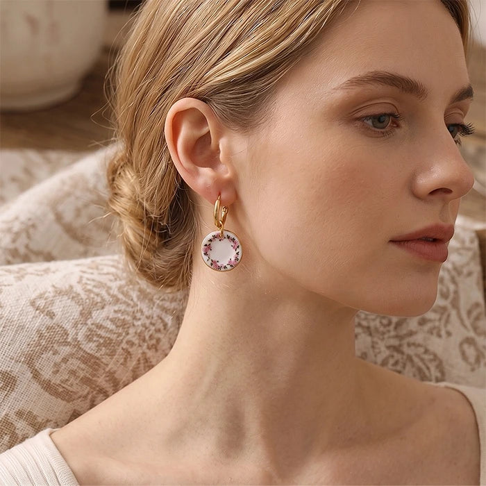 A woman wearing a gold hoop earring with a circular drop featuring a pink and white floral pattern, shown in a close-up profile view.