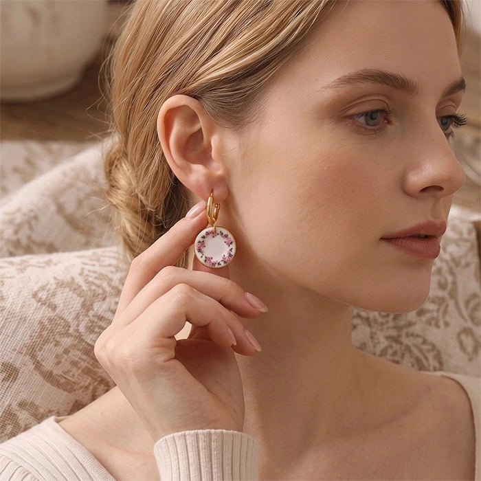 A close-up lifestyle shot of a woman wearing a gold hoop earring with a circular white and pink floral pendant, holding the earring with her hand.
