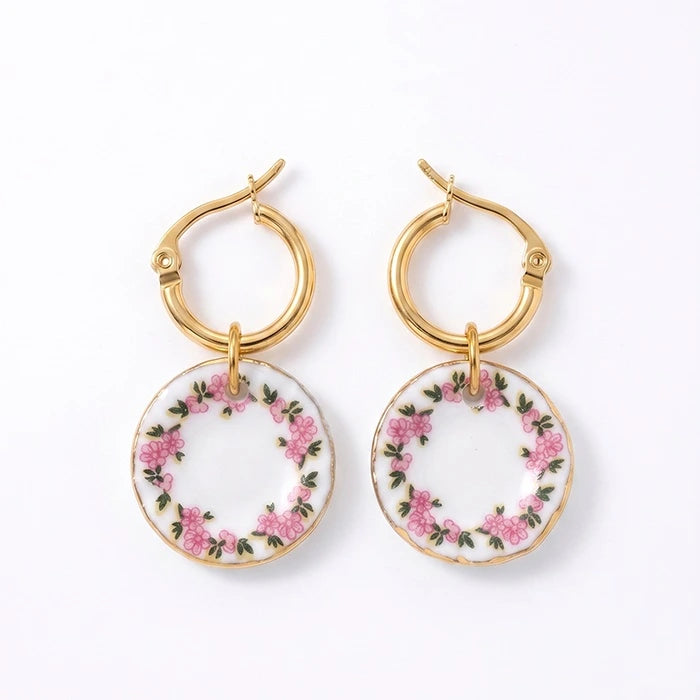 A pair of gold hoop earrings with round white porcelain dangles decorated with a pink rose and green leaf floral wreath, shown on a clean white background.