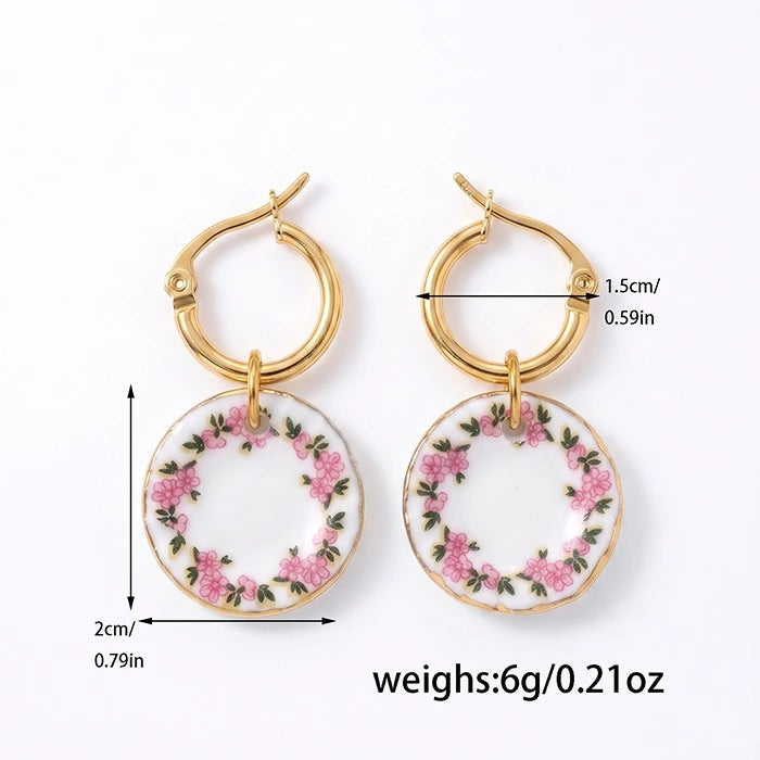 Gold hoop drop earrings with circular white porcelain pendants decorated with pink roses and green leaves, shown with dimension labels and weight text on a white background.