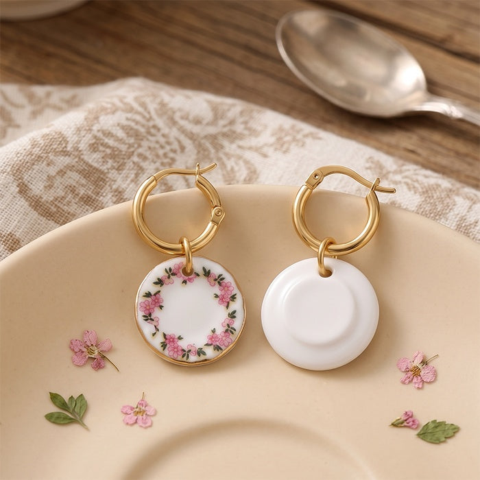 A pair of gold hoop earrings with round white ceramic charms, one decorated with pink floral patterns and one plain white, displayed on a beige plate.