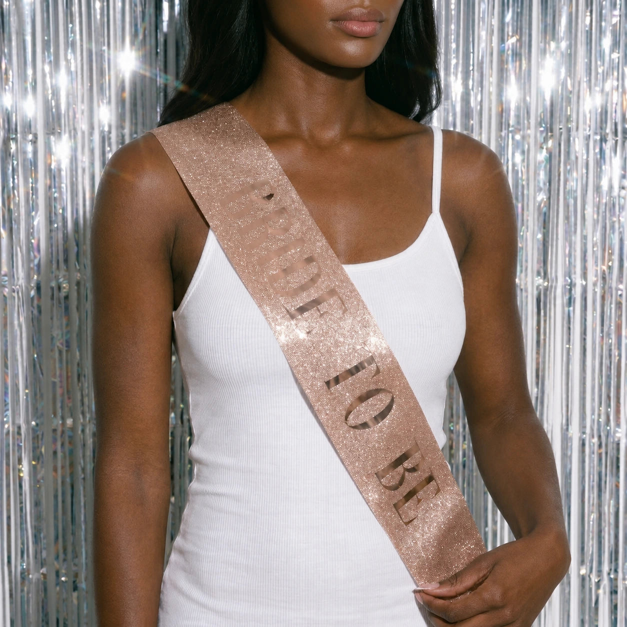 Woman wearing a white dress with a rose gold 'Bride to Be' sash against a silver tinsel background