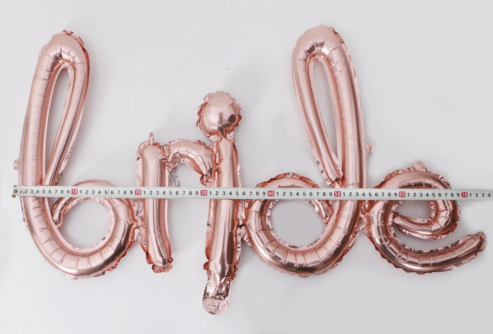 Rose gold foil balloon letters spelling 'Bride' laid out on a white background, with a measuring tape stretched horizontally across the balloons to show scale.