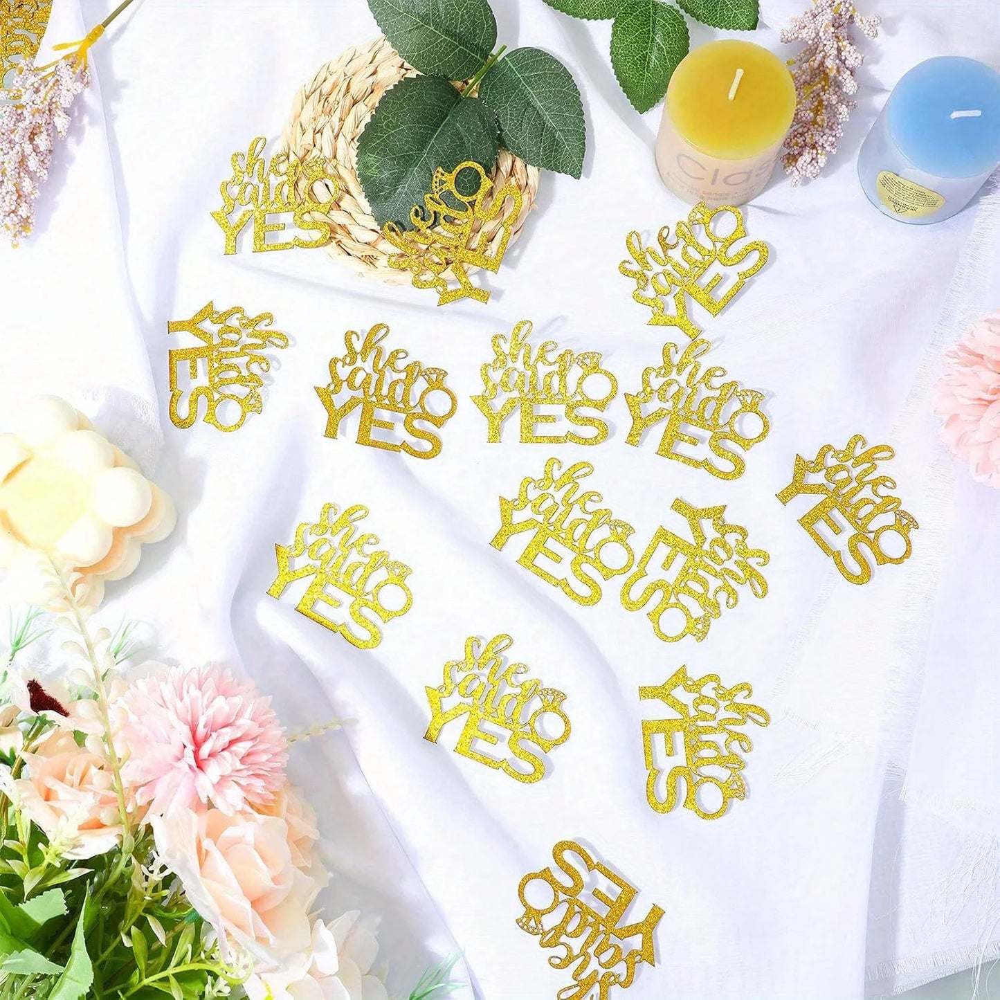 Gold 'She Said Yes' confetti on a white surface with flowers and candles.