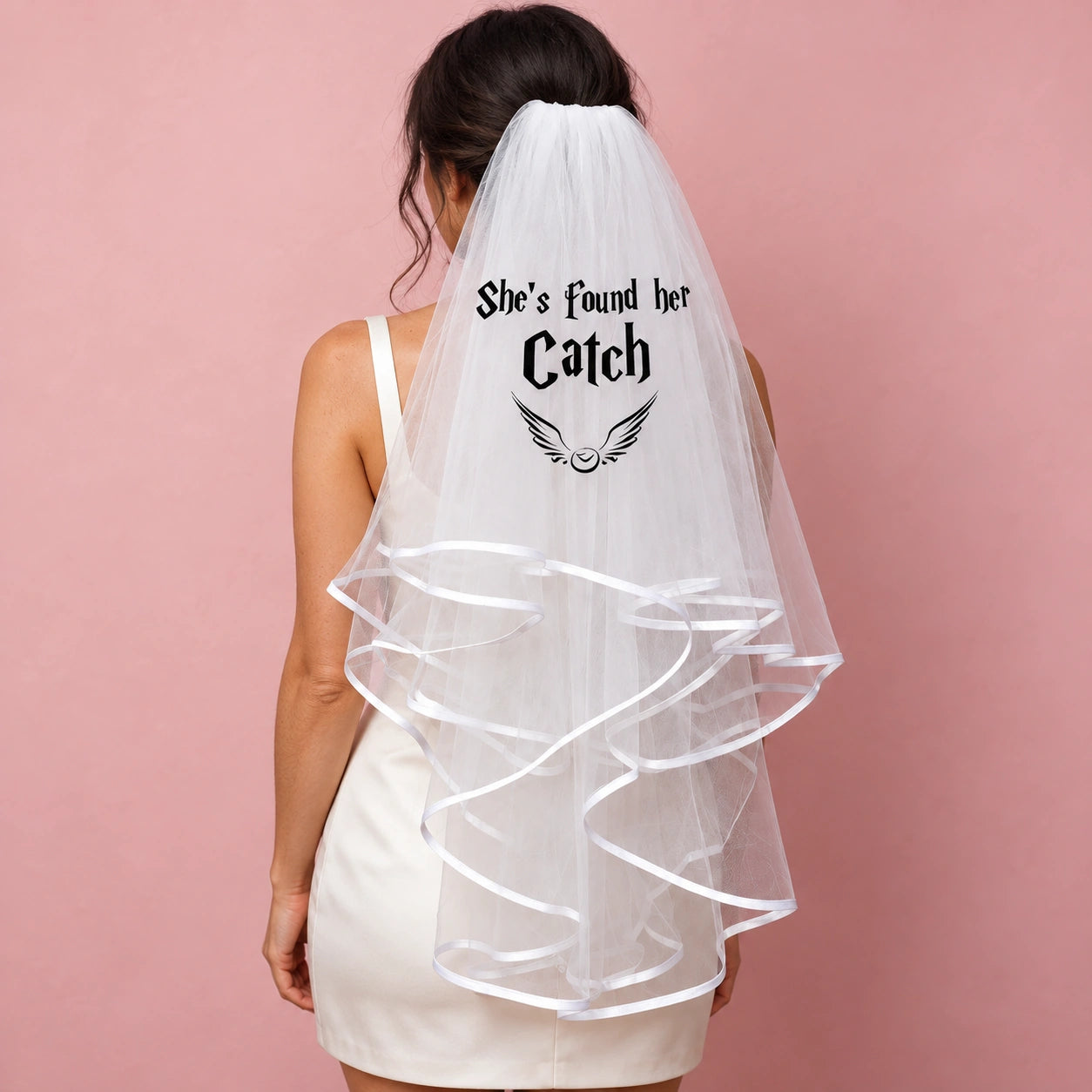 A woman shown from the back wearing a white bridal-style veil with black printed text reading 'She's found her Catch' and a small wing graphic, against a solid pink background.