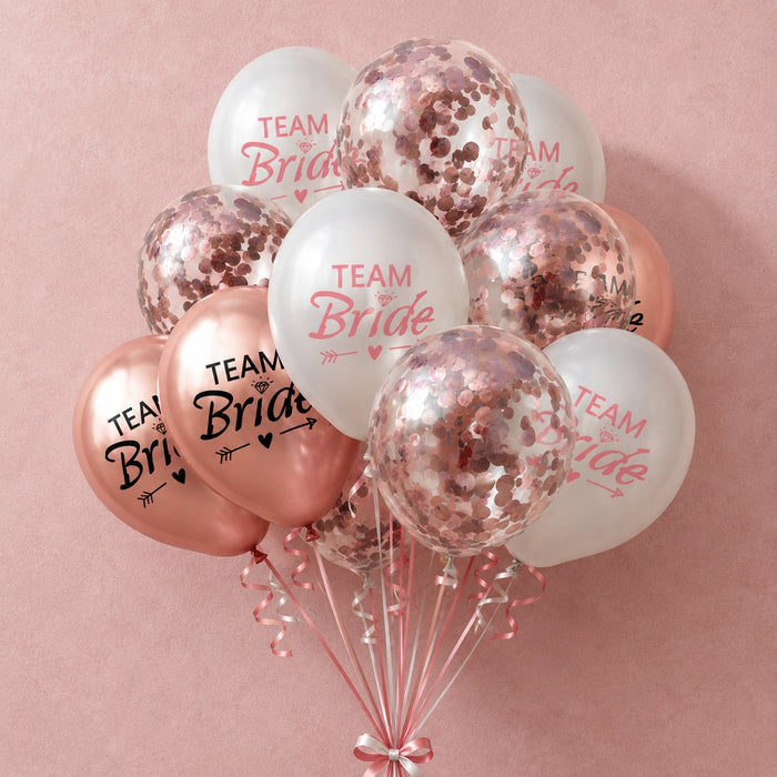 A cluster of bridal shower balloons on a dusty pink background. The set includes white latex balloons printed with 'TEAM Bride' in pink script, rose gold foil balloons with 'TEAM Bride' in black, and clear balloons filled with rose gold confetti.