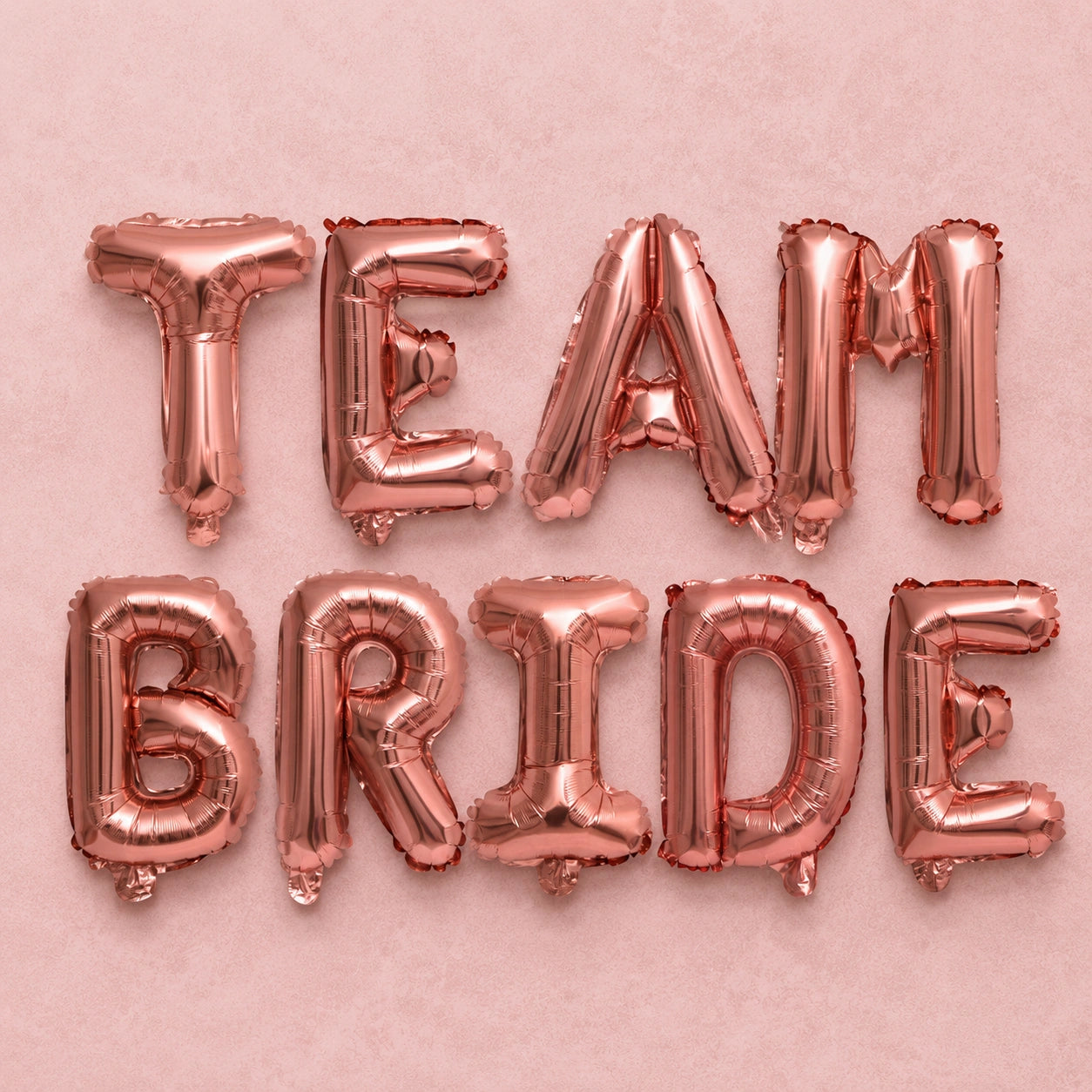 Rose gold foil balloon letters spelling out 'TEAM BRIDE' arranged in two rows on a textured pink background.
