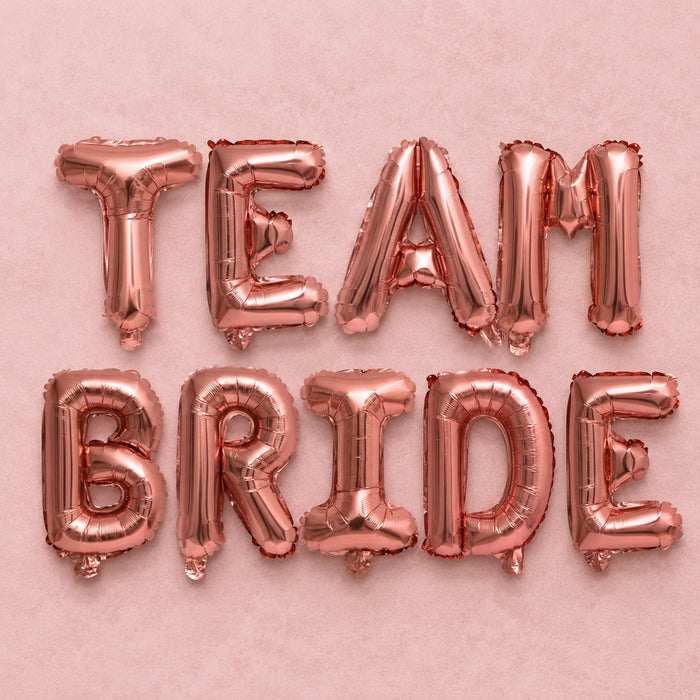 Rose gold foil balloon letters spelling out 'TEAM BRIDE' arranged in two rows on a textured pink background.