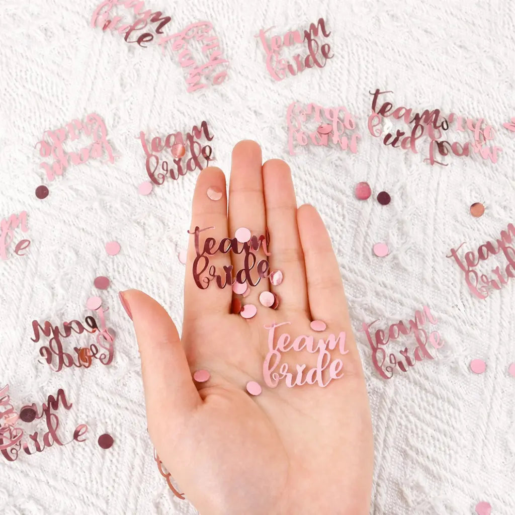 Hand holding 'Team Bride' confetti with more confetti scattered on a white background