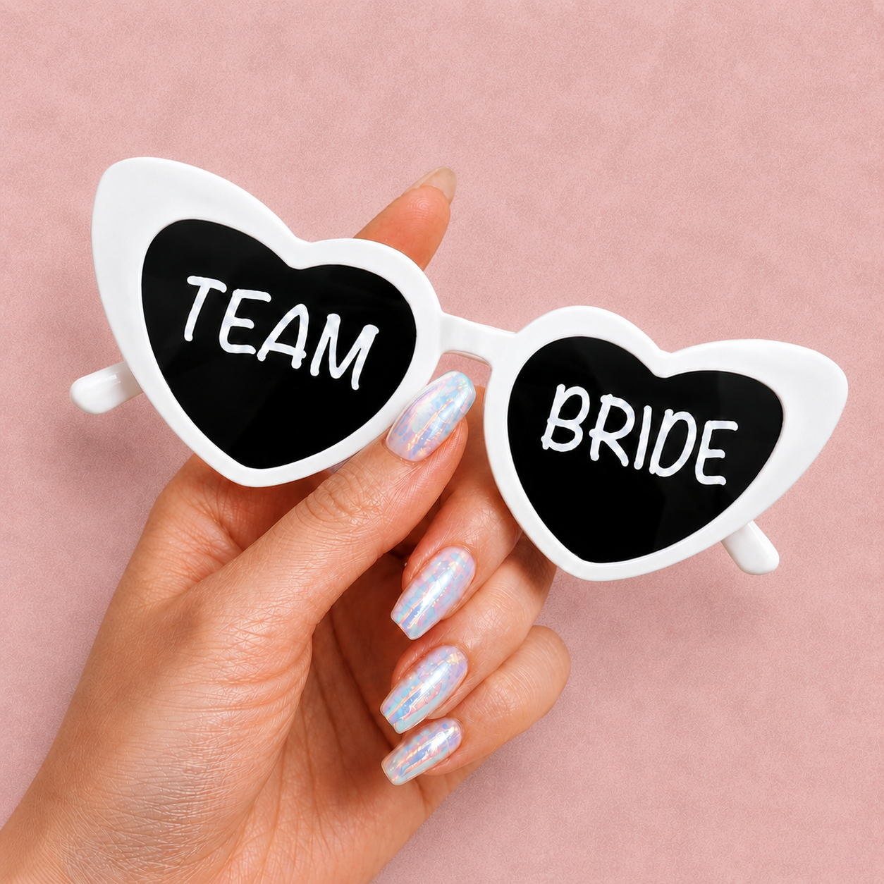 Heart-shaped sunglasses with 'Team Bride' text held by a hand on a pink background