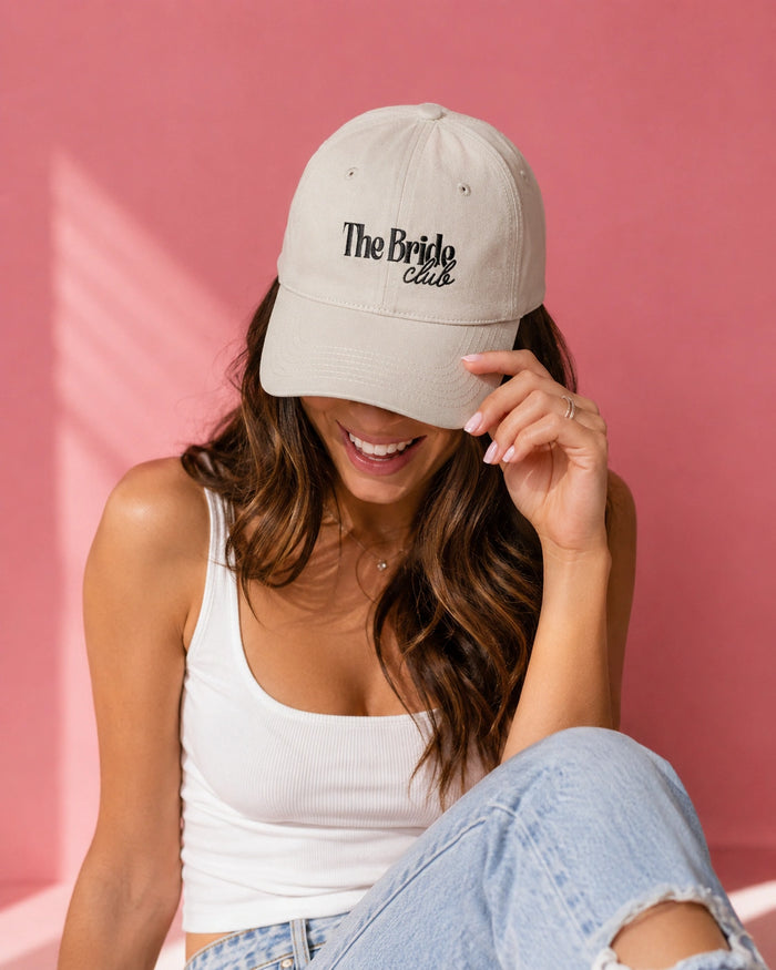 A woman wearing a beige baseball cap with 'The Bride Club' embroidered in black on the front, posing against a solid pink wall.
