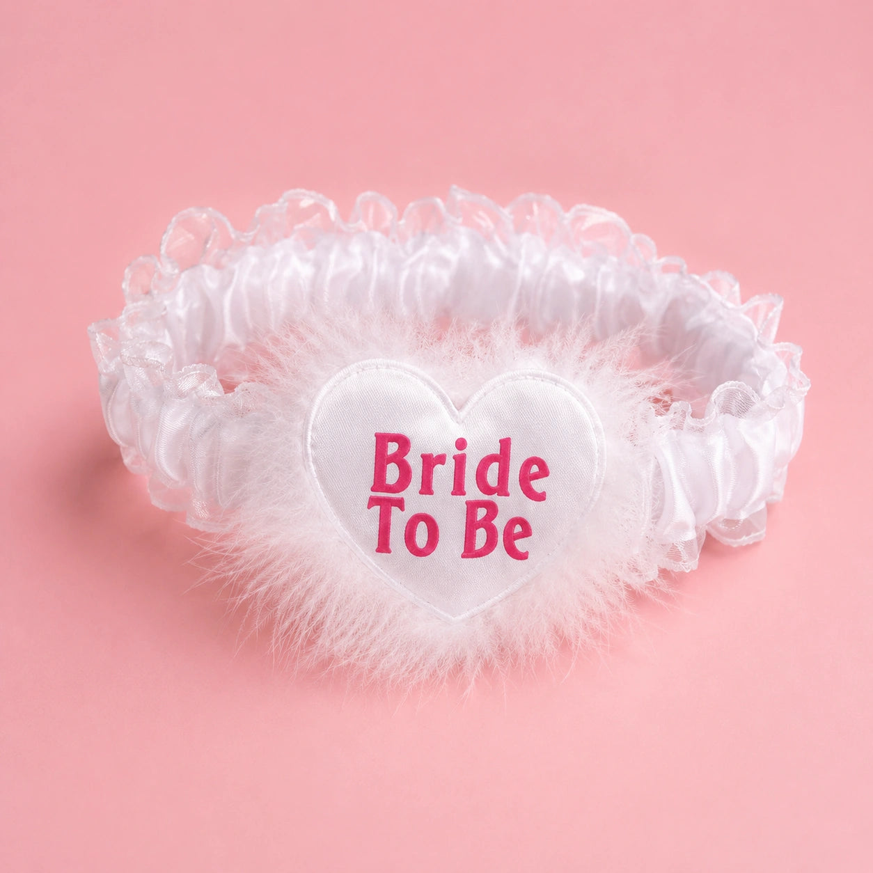 A white bridal garter with a ruffled satin band, fluffy tulle trim, and a central white heart patch with pink text reading 'Bride To Be', set against a solid pink background.