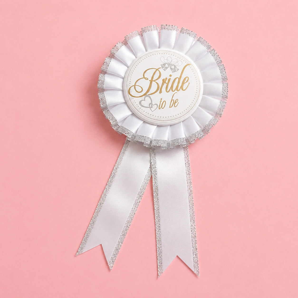 A white bridal rosette badge with silver glitter trim and a white ribbon tail, featuring a central round label that reads 'Bride to be' in gold script, set against a solid pink background.