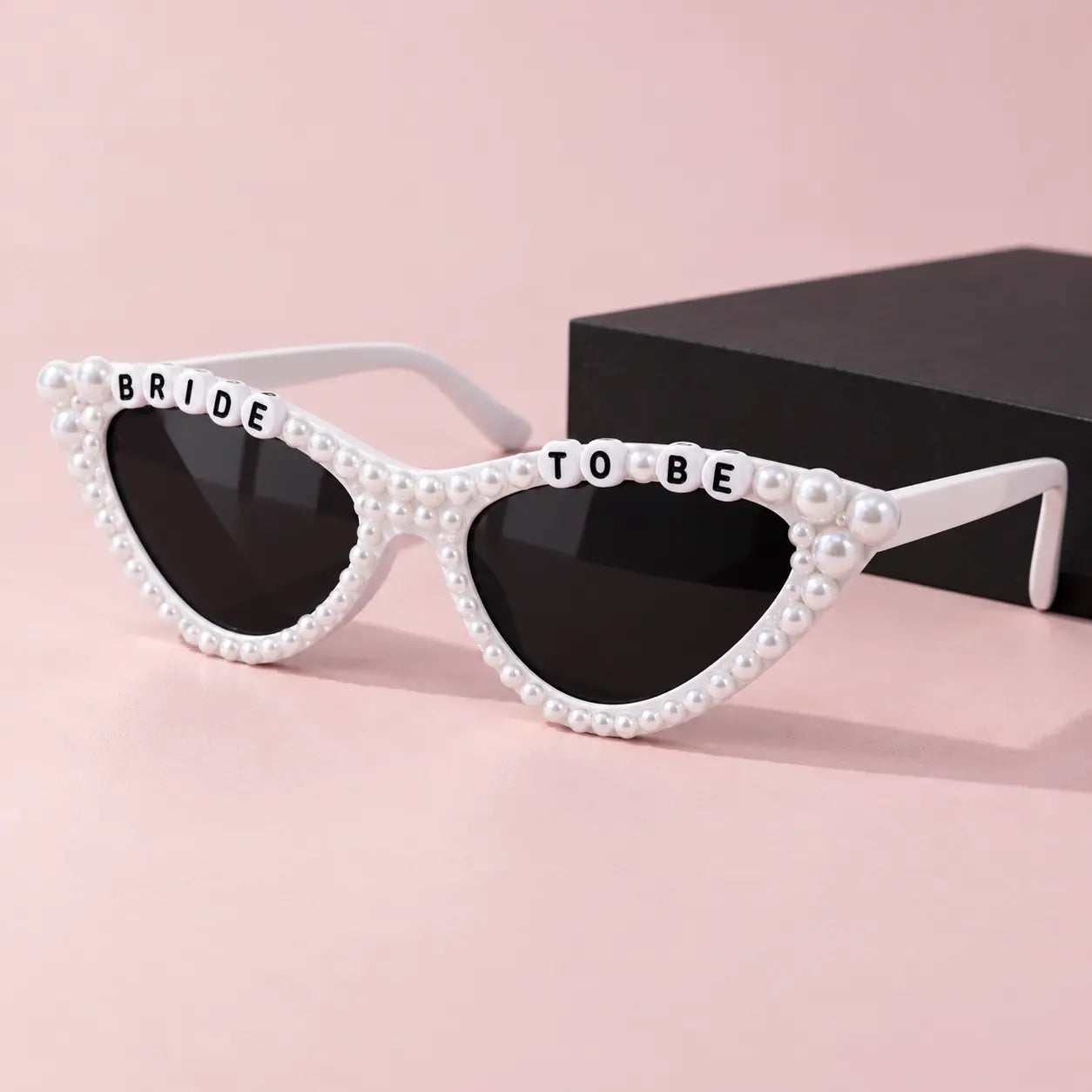 White cat-eye sunglasses embellished with faux pearls and letter tiles spelling 'BRIDE TO BE', shown on a soft pink background with a black box behind them.