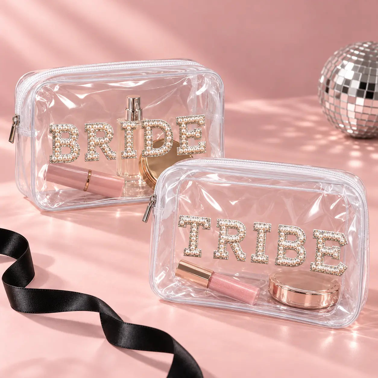 Two clear zip cosmetic bags on a pink studio background. The front bag says "TRIBE" in rhinestone letters and contains a pink lip product and rose gold compact.