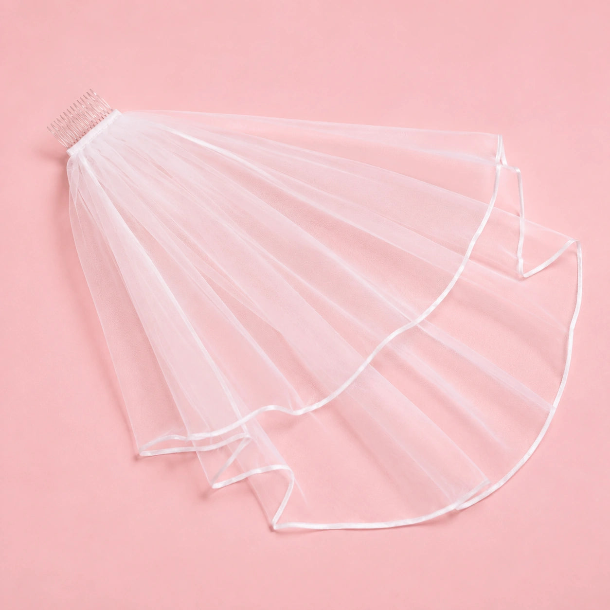 A white tulle bridal veil with a built-in hair comb, laid flat on a solid pink background.