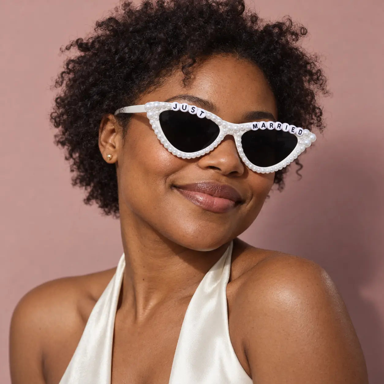 A woman with curly black hair wearing white rhinestone-trimmed cat-eye sunglasses that say 'JUST MARRIED' on the bridge, posed against a dusty pink background.