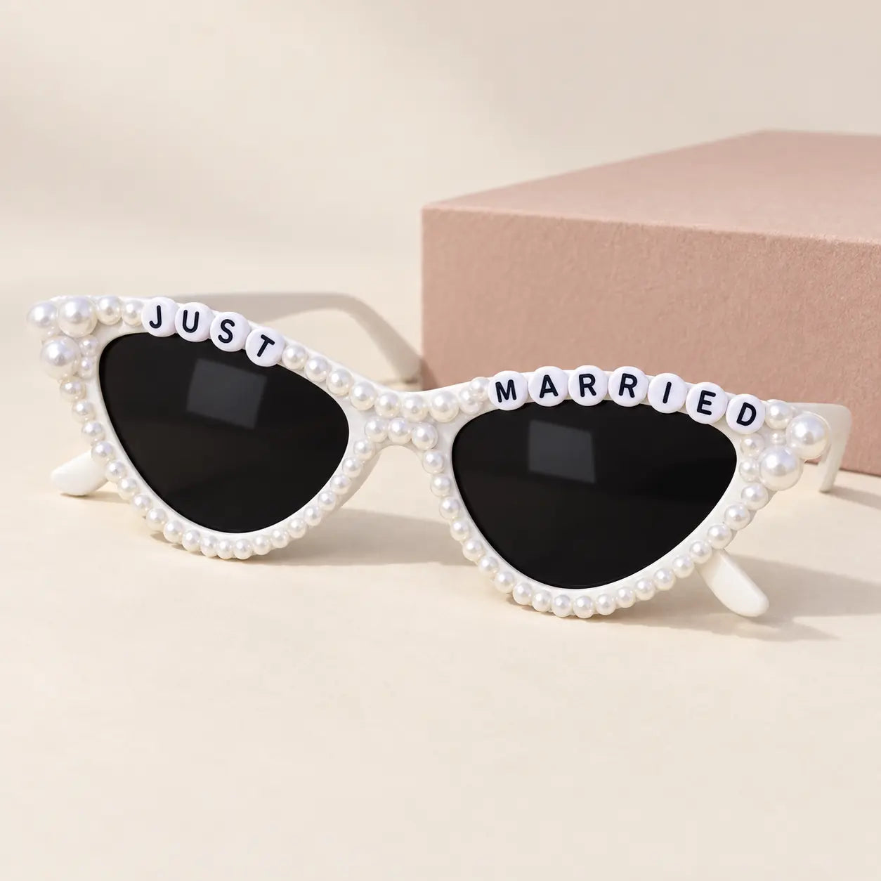 White cat-eye sunglasses embellished with faux pearls and letter tiles spelling 'JUST MARRIED' across the top, shown on a warm beige background with a pink box behind them.