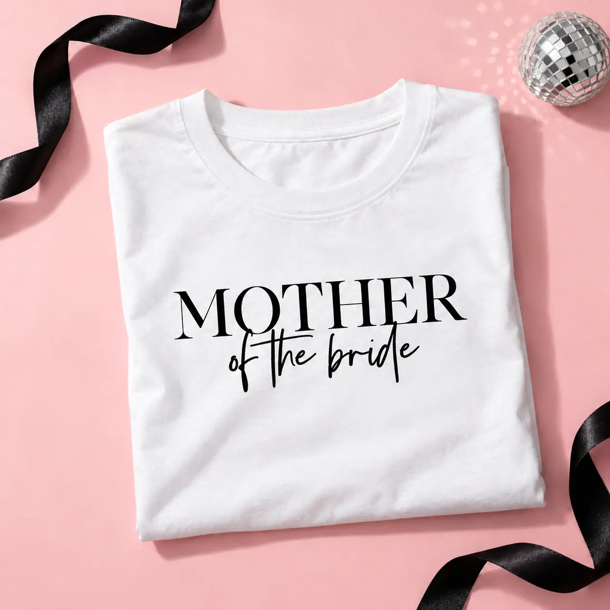 A folded white t-shirt with the text 'MOTHER of the bride' printed in black, styled on a pink background with black ribbons and a disco ball prop.