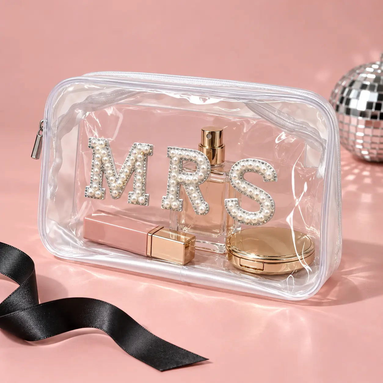 A clear zippered cosmetic pouch with the letters 'MRS' embellished with rhinestones, containing a perfume bottle, lipstick, and compact.