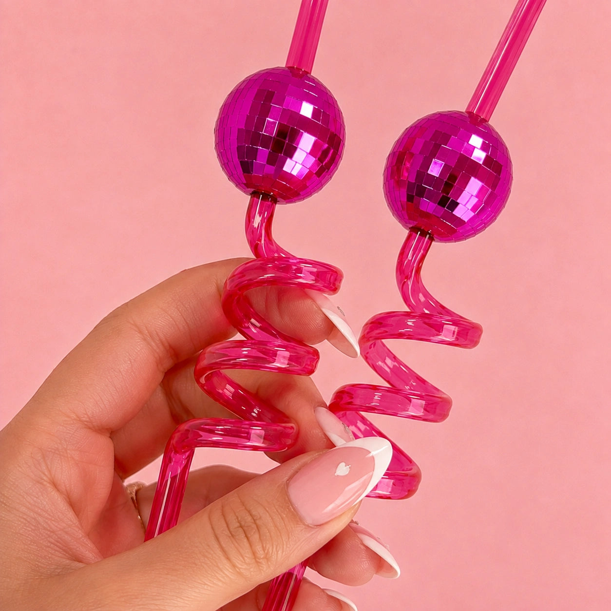 A hand with a French manicure holds two bright pink spiral straws with disco ball-shaped tops against a solid pink background.